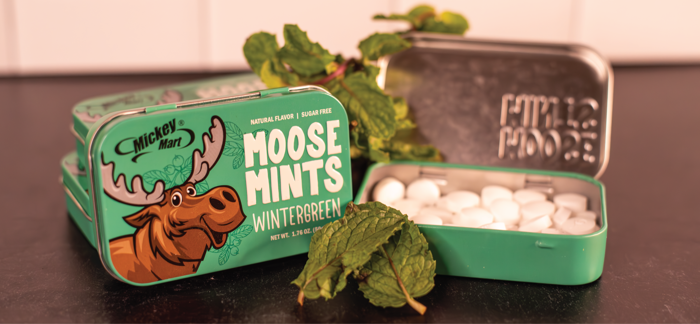 Metal Mint containers with one open showing the small white mints inside. The tin cover has a moose and the words "Moose Mints - Evergreen" and a few mint leaves rest on the stack of tins